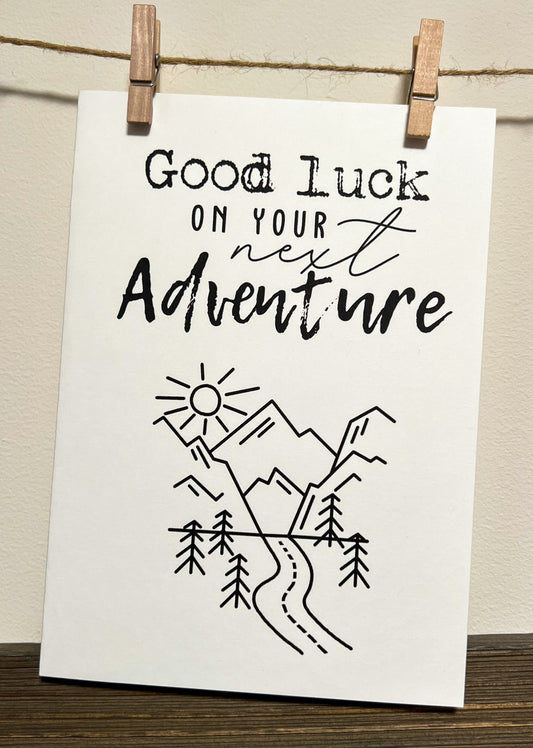 While cardstock card with black writing ' Good luck on your next Adventure' with a photo of a road going through the mountains with a sun and trees. Blank Inside.